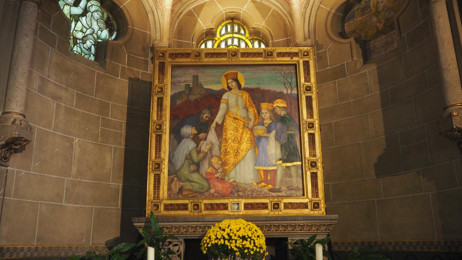 Elisabeth-Altar von Matthäus Schiestl. Der Elisabeth-Altar im nördlichen Seitenchor entstand 1915.  Er zeigt die Markgräfin von Thüringen Elisabeth, wie sie mit ihren beiden Kindern Brot und Kleidung an die Armen verteilt.  Elisabeth von Thüringen (* 1207[1] in Pressburg oder auf Burg Rákóczi in Sárospatak (Ungarn); † 17. November 1231 in Marburg an der Lahn), auch Elisabeth von Ungarn genannt, war eine ungarische Prinzessin, deutsche Landgräfin, ist Heilige der katholischen Kirche und galt als „deutsche Nationalheilige“. Ex-Bild-DB-ID: 27078